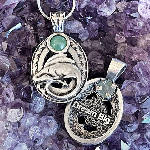 Silver pendant laying on a bed of amethyst. The pendant features a sleeping dragon with an aventurine moon hovering over it. The back of the pendant says "Dream Big".