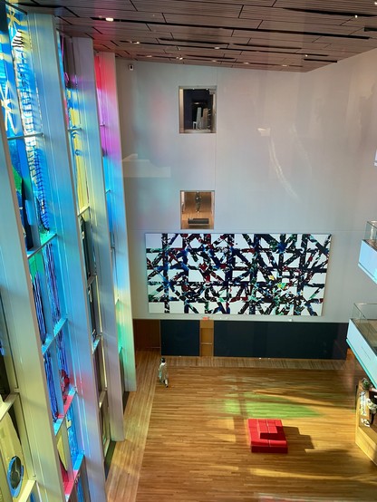 A shot of the lobby of Charlotte’s Mint Museum from the 4th floor. Sunlight is shining through stained glass windows. There is a modern abstract painting, wood floors, and a red couch.