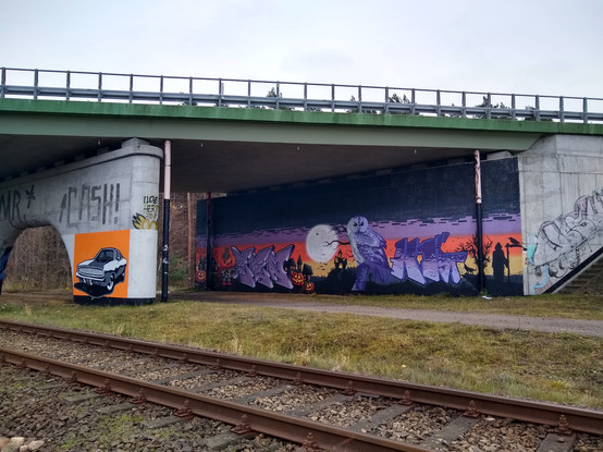 A viaduct behind a railway line, with road on top. A large Halloween-themed street art covers the inside. There is also a small art of a old-school sports car on an orange background painted on the bridge support in the front.

The Halloween-themed painting features a number of figures on a sunset-colored background (but without the sun). The figures include, left to right, a jack-o-lantern in front of a spooky looking house, a boy with a pumpkin head, some nondescript purple letters, another spooky house with two more jack-o-lanterns, a huge white moon with bats in front, one more spooky house on a hill, a huge purple owl, more purple letters, a howling wolf besides a bare tree, and a grim reaper with a scythe and a crow perched on its blade. The background consists of bands of yellow, orange, purple and then black. Wide black rectangles are randomly spread on top of the purple band.