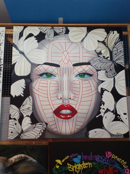 a painting of an ivory-colored woman with red lips and a red minimalist line design on her face and green eyes looking at the viewer, the painting is of just her face, she has dark hair and her face is encircled by large butterflies