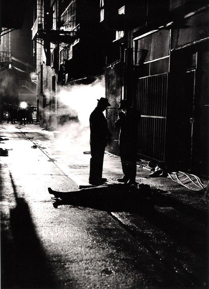 two silhouetted men in a dark smoky street, another lies prone on the ground