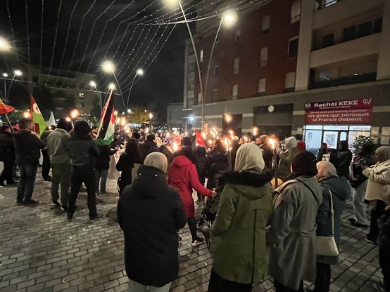 rassemblement sur une place en ville. On voit des personnes debout, certaines avec des flambeaux, on voit quelques drapeaux : Palestinien, du PG, etc.
on voit la devanture de la permanence parlementaire de Rachel Keke.