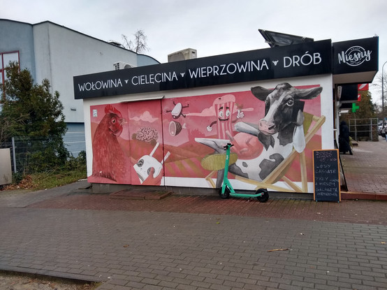 A mural on a meat shop. The label above says "beef, veal, pork, poultry".

The mural depicts anthropomorphized animals on a red-tinted background of hills. The hills are covered by alternating stripes of green and red.

On the left side, there's a huge hen with human hands, holding a gamepad-style drone controller. The drone is seen flying away, carrying a large piece of ham on strings.

On the right side, a cow is sitting on its back, on a deckchair. Its left front hoof is holding a phone in front of its eye. Its right front hoof is laid across its udder. The right back leg is crossed on top of the left back leg and it is wearing some kind of shoe.

Behind the cow, a striped piece of meat is seen standing. It has a smiling mouth, two eyes with brows and two stick-hands finished with white palms. The left hand is either showing a thumbs-up gesture or holding something, possibly both.

In the distance, a huge brain is seen lying on the hill in the left. A show of a building — possibly a factory — is seen in the middle.