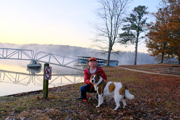 A man with an orange cap crouching, while holding his white and brown dog. A lake with steam rising from water, trees on the right top, a bridge to a deck, and a boathouse in the background