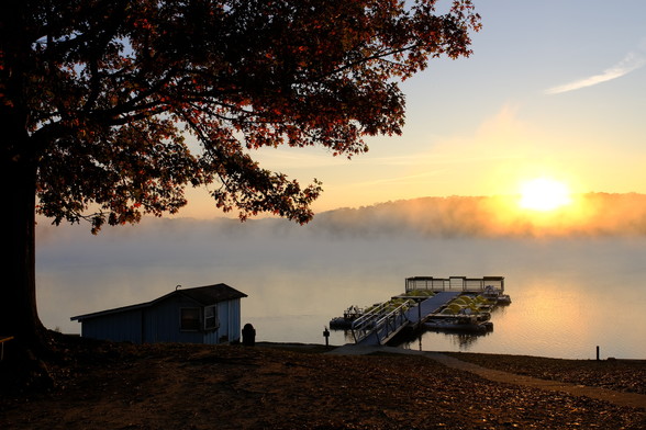 A lake view with the sun just rising over a hill, tree branches and leaves hanging over it from left top, a boathouse on bottom left, and a deck with paddle boats on the side