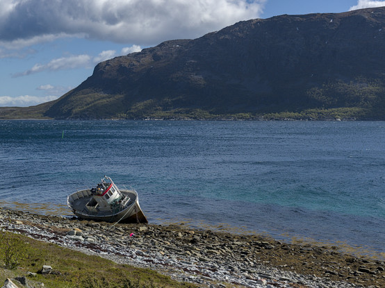 The pictures shows a fishing boat lying on the side at the beach at low tide. In the back you see a mountain range on the other side of the bay. The sky is blue and with some clouds. The small waves on the water show that there is some wind blowing.