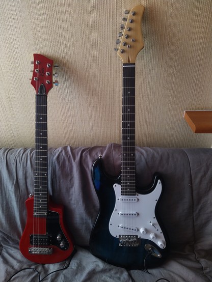 Foto donde aparecen dos guitarras electricas sobre un sofá. La guitarra que esta en el lado izquierdo es de color rojo, la guitarra de la derecha es de color verde y blanco.