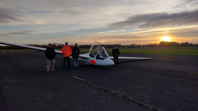 A glider on the runway, with the setting sun behind, and several ground crew, plus pilots, smiling for the camera