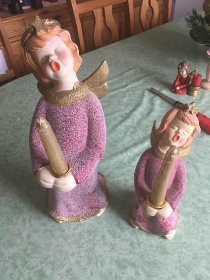Two angelic candleholders are shown holding tapered candles in their hands. Each (female) angel has golden hair and is wearing a plum-colored (choir?) gown with a golden scarf around their necks, and each have their pale faces upturned with open mouths to suggest they are singing. The candles are also golden and glittery except for where the white wick pokes out of the smooth, glitterless wax at the very top. The angels are casually holding the candles so that the tops of the candles are pointing directly at their open mouths, suggesting that the candles are penises about to be fellated. 😲