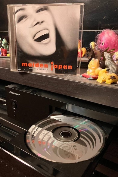 Jewel case with cover. Black and white photo of a laughing woman. "Maiden Japan" in block orange letters at the bottom and Monday Michiru in grey. Below is a silver CD with grey circles in a CD player tray.