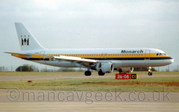 Side view of a white twin engined jet airliner with a yellow and black stripe running along the body and black "Monarch" titles on the upper forward fuselage, taxiing from left to right., under a flat grey sky.