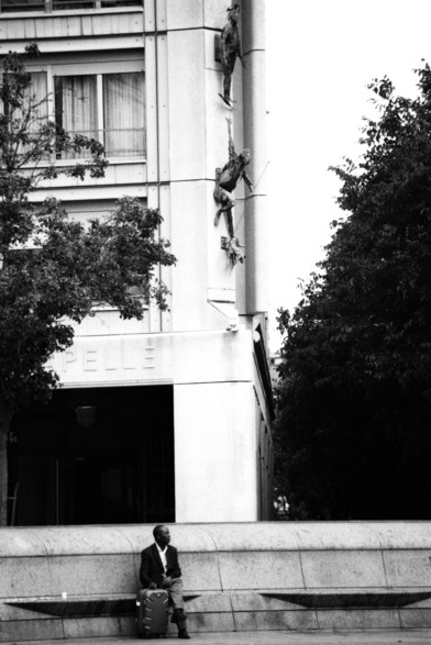 Homme assis sur un banc au pied de l'angle d'un immeuble, noir et blanc.