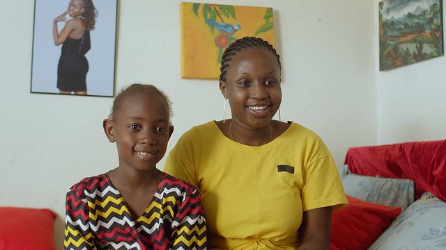 A young woman in a yellow dress and a young girl in a print african dress in a scene in a film, staring at something off screen. White walls in the background with a few photos and paintings on it.