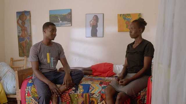 A scene in a TV series produced in Gulu, Uganda. Two actors sit on a bed facing each other, male and female, the walls are white with light from the window creating patterns.