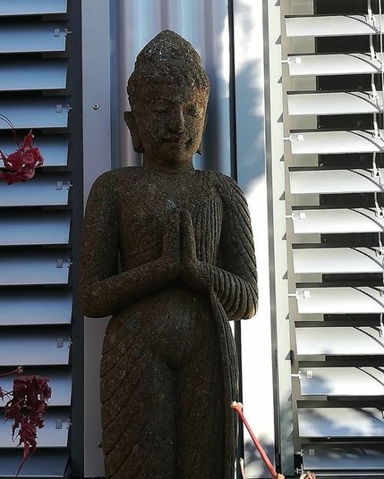 Stehender Buddha mit gefalteten Händen. Im Hintergrund sind Lamellen einer Fensterjalousie zu sehen.