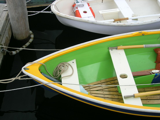 Closeup view of a green rowboat floating in dark water tied to a dock. A smaller white rowboat floats next to it.