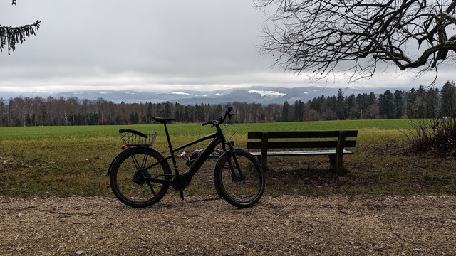 An eBike and a bench in front of a field. Woods in the back, and then snow-covered hills. Grey sky. It looks cold.