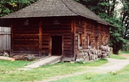 A working post and sill building, not whitewashed, but with shingle roof. This is the blacksmith'sshop and cooperage, as you can see from the firewood and barrels stacked along the side of the building.