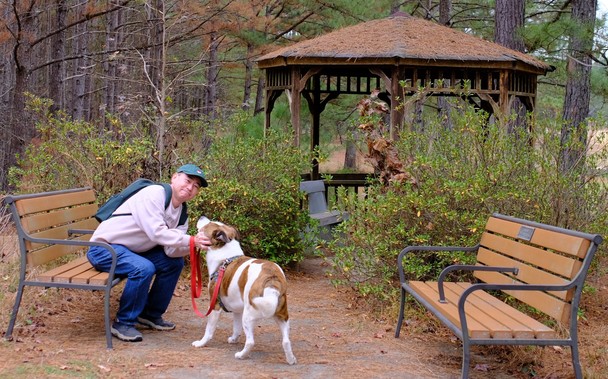 A man with a green hat and a backpack sitting on a park bench holding his white and brown dog. A gazebo is in the background.