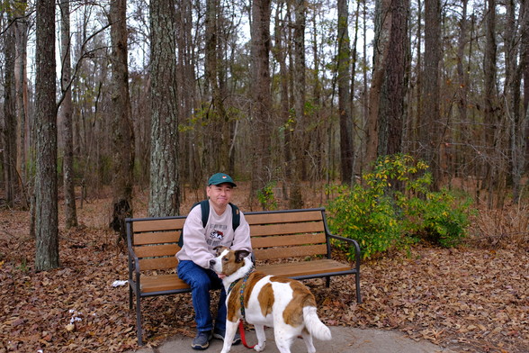 A man with a green cap sitting on a park bench holding his white and brown dog. Trees behind.