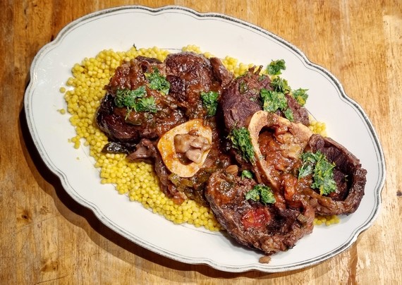 Eine weiße ovale Platte, darauf gelber Couscous, geschmorte Rinderbeinscheiben, dunkle Sauce und eine grünliche Gremolata