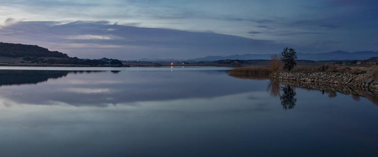The image shows a reservoir with calm water and low light (during the blue hour). You can see some trees at your right and some low mountains at your left.