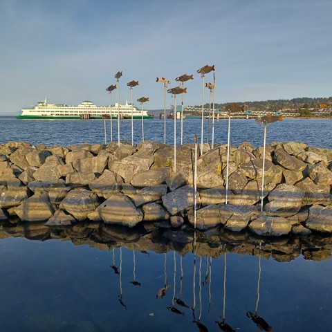 Photo of metal salmon atop long poles that have been securely attached to the rocks near the dock. There is a large green and white ferry in the background, blue sky with a few clouds, calm water.
