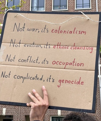 A piece of cardboard being held up by a hand, written on the cardboard: Not war, it's colonialism. Not eviction, it's ethnic cleansing. Not conflict, it's occupation. Not complicated, it's genocide.