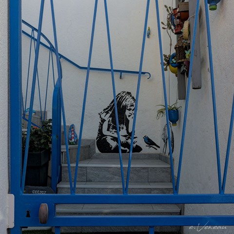 A house entrance, the bottom of a staircase, a metal gate painted blue. Behind on a white wall, at the bottom of the stairs, the stencil of a little girl, chin in her hand, looking at a bird with blue feathers on a branch.