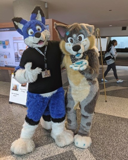 Nev, a blue fox fursuiter and mokie, a doggo fursuiter pose for a photo together