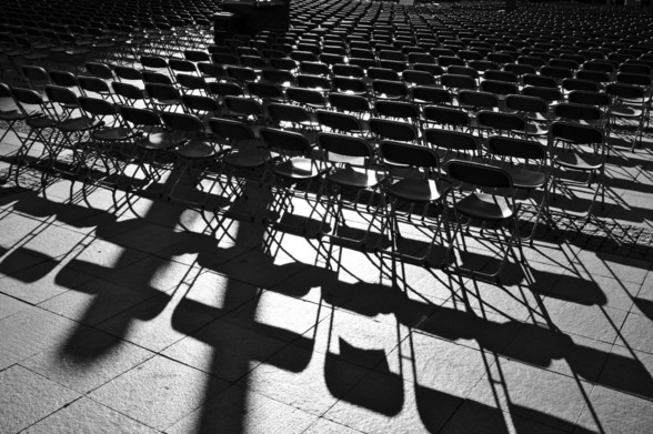 SchwarzWeiß Bild von eng gestellten Stuhlreihen vor einer Open-Air-Tribüne im Gegenlicht. Dramatische Schatten im Vordergrund