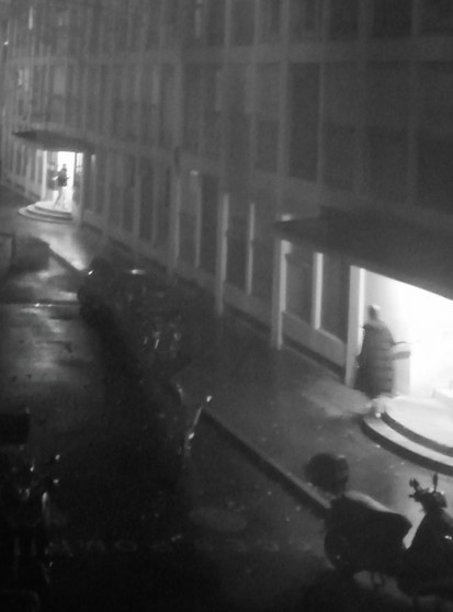 Exterior housing project building, evening, A misty blurry rainy greyish evening (b and w photo);   two entrances to the buidling are lit, in the first one on a step a woman's silhouette looking toward   the second entrance thirty meters away and the two silhouettes on the steps.