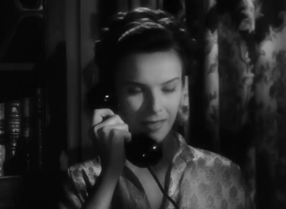 A woman is holding the phone. Her hair is tied. We can see a curtain behind her.