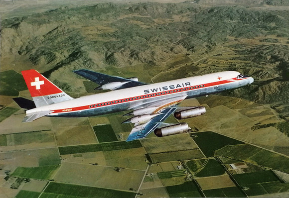 4 engine jetliner flies over green square farm land and small mountains . Perspective is looking down and to the left over the wings and fuselage