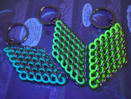 three glow in the dark keychains in the shape of a diamond. they are made with the dragonscale chainmaille weave and come in blue, green, and yellow for the glow rings, which are paired with stainless steel rings.