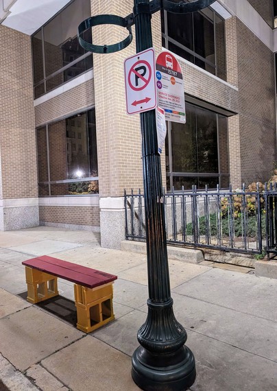 A photo of a red and yellow bench made of cinderblocks next to the Euclid @ BJC bus stop in St. Louis, Missouri. The bench is on the sidewalk next to the stop and there is a building alcove and a black iron fence in the background.