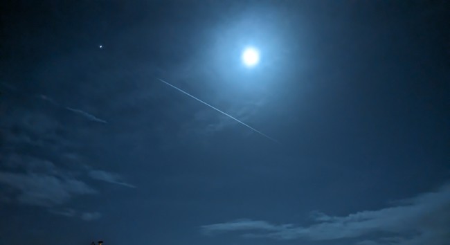 Photo of a blueish night with sparse clouds, ice crystals and a jet trail framing the moon.