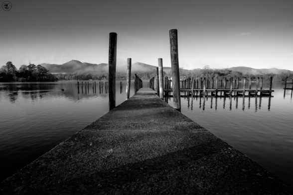Monochrome shot along concrete pier with raised wooden stakes. Still lake waters surrounding, with mountains covered in mist in background