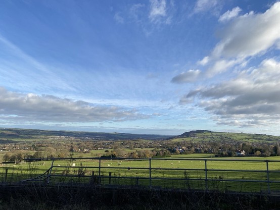 Looking over a fence and a field of sheep to hills beyond. There are white fluffy clouds in the sky.