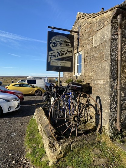 Bicycles parked between two rusty cartwheels in front of a stone building and under a sign that reads “Tan Hill Inn - Britain’s Highest Inn, 1732ft ASL”