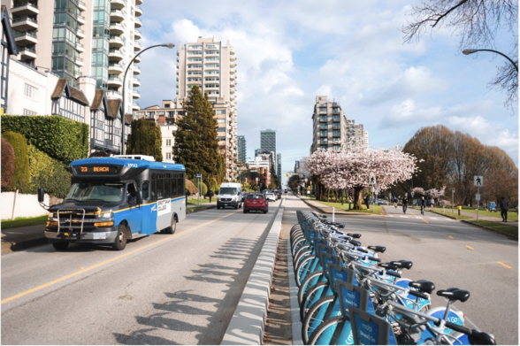 A small capacity blue and grey bus on the street with an array of bikes for hire and some other traffic. A cherry tree is in flower and the sky of Vancouver's West End is partly cloudy. There are several tall residential buildings