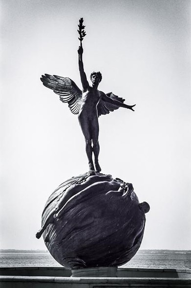 Statue of a naked angel figure reaching up with a frond in outstretched arm.  Wings spread out behind. standing of a globe which has overlapping female limbs on it.
