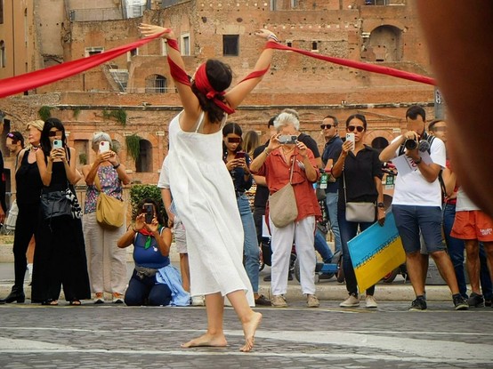 an iranian woman holding a dance performance on the street  at the march i mentioned in the caption .  many people are taking pictures/videos around her .   unfortunately i don't know her name ,  please let me know if you recognize her.
  she's got medium/long  dark hair ,  she's  dressed in white and barefoot.   she's gracefully dancing  using  a very long red tape ,  that is  tied to her head  and also  held by other assistants  (but  you can't see them in these pictures).    right now ,  she's turned around  while holding up her arms  that are wrapped in said tapes .   her eyes are covered by the tape as well
