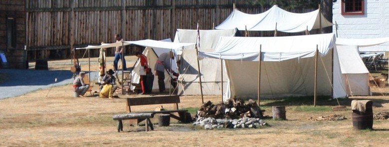 typical encampment of the fur trade, with canvas tents for the gentlemen and cook fires for all. To the left men talk under a canvas fly. In the background the palisades of replica Fort Langley.
