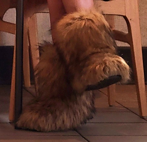 A person's legs crossed with big boots over the feet. The boots have shaggy brown outer layer like a yak's hair. The person is sitting on a brown wooden chair though that detail is not visible.