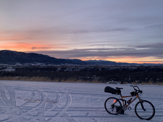 High pink and red sunset clouds over the Beartooth mountains seen through clear winter air with an orange bicycle on a snow covered gravel road in the foreground.