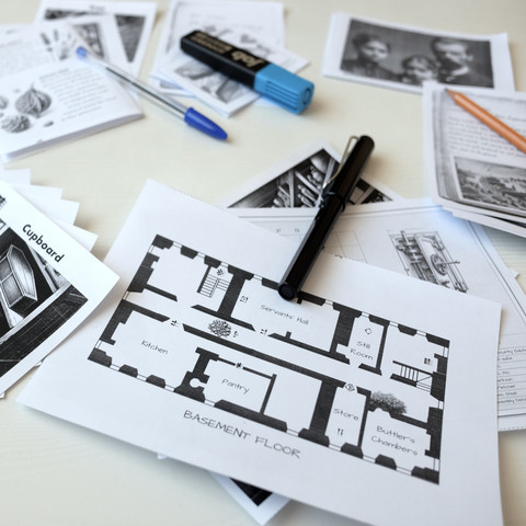 Printed components of the second episode of 'The Secret of Langton Manor' print-and-play escape room game on a desk, featuring puzzle pages, a detailed basement floor plan, photos, notes, and a couple of markers.