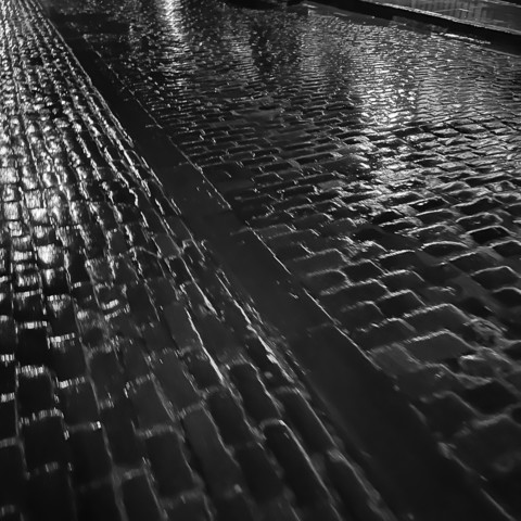 Square monochrome photo.
A wet road, made from stone setts, gleaming in reflected lights (not visible).
Larger stones form a kerb, running diagonally from the bottom right. Setts to the left are laid with the long edge vertically, whereas the road to the right has them laid long edges across the photo.