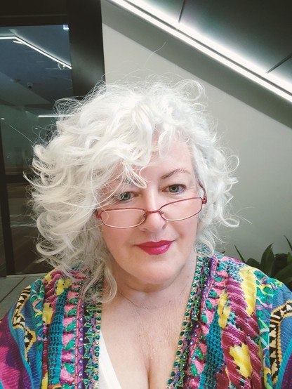 A woman with white curly hair and a colourful top wears Barbie core lipstick and cute reading glasses.