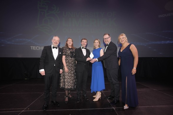 A photograph of six people in smart evening wear on stage at an awards event. They are gathered around a trophy, with a large screen behind them displaying the University of Limerick logo